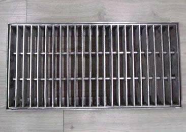 A swage-locked drainage trench box grate is installed on the wood floor.