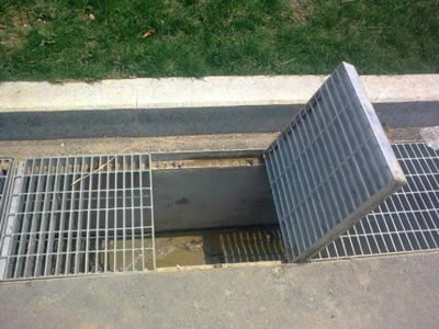 Several GM type steel grating trench covers are installed edge of road with one grating opened.